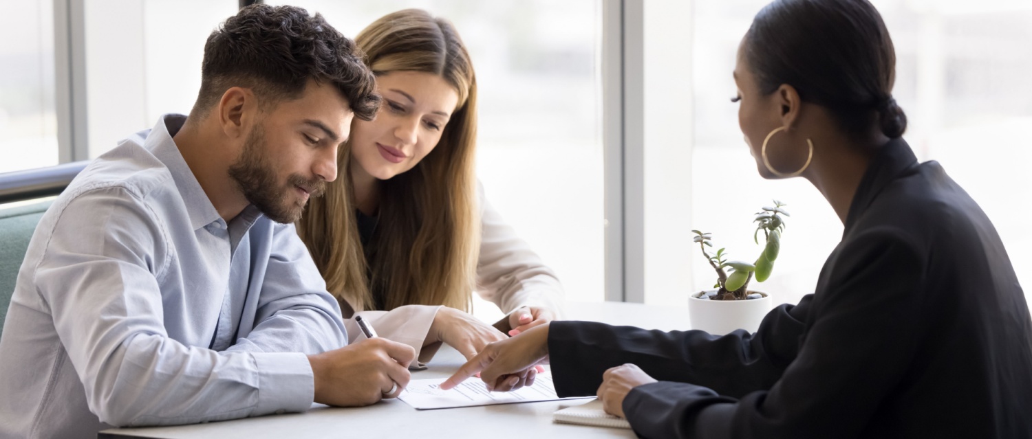 Confident young African realtor agent pointing at paper agreement for couple of customers, clients signing, giving consultation, legal help with mortgage, real estate property buying