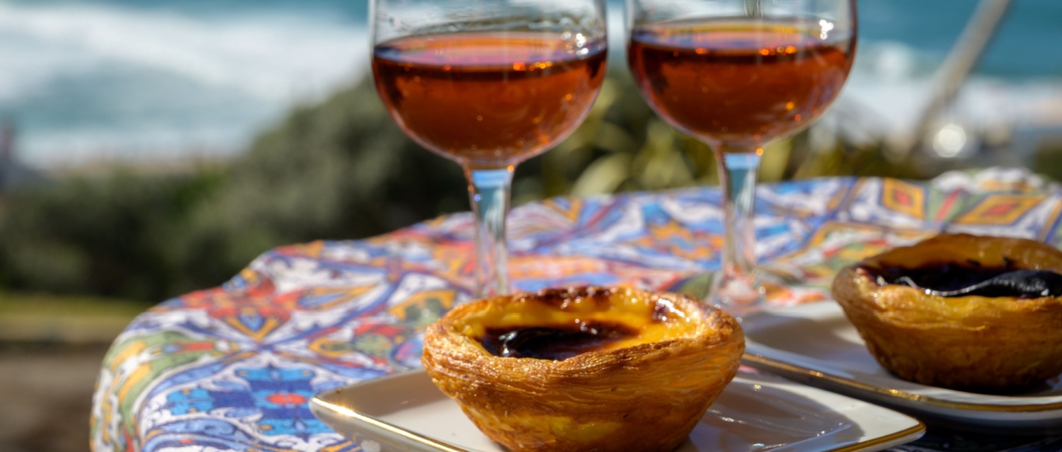 Portugal's traditional food and drink, glasses of porto wine or muscatel de setubal, sweet dessert Pastel de nata egg custard tart pastry served with view on blue Atlantic ocean near Sintra in Lisbon